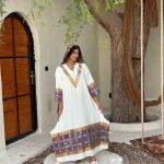 زنی با لباس بلند سفید و حاشیه‌های طرح‌دار رنگارنگ، کنار درخت و تاب در حیاط ایستاده است